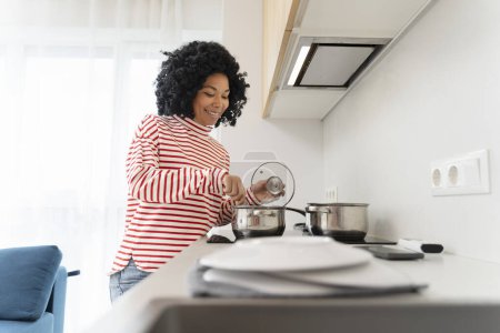 Foto de Retrato de una hermosa mujer afroamericana sonriente cocinando en una elegante cocina moderna, sosteniendo la cacerola, en casa en el apartamento. Ama de casa positiva usando equipo y platos. Concepto de hogar - Imagen libre de derechos