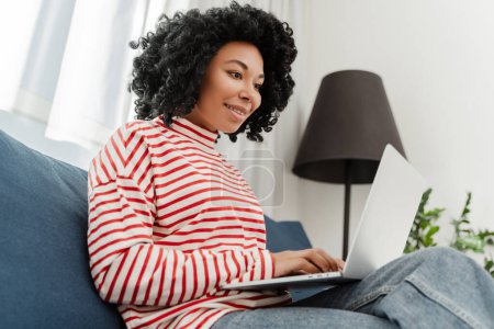 Téléchargez les photos : Belle femme afro-américaine souriante, pigiste utilisant un ordinateur portable travaillant en ligne à partir de la maison - en image libre de droit