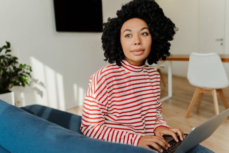 Téléchargez les photos : Portrait de la femme afro-américaine bouclée réussie assise sur un canapé confortable à l'aide d'un ordinateur portable, dans un appartement moderne. Belle femme portant des vêtements décontractés travaillant en ligne, les médias sociaux - en image libre de droit