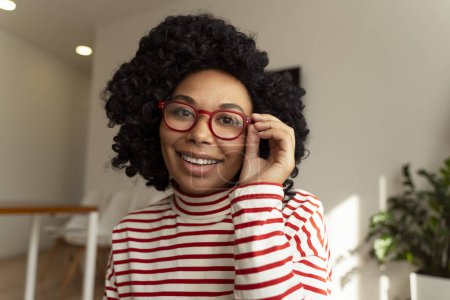 Foto de Retrato de una atractiva mujer afroamericana sonriente con elegantes gafas rojas, mirando a la cámara. Hermosa mujer en ropa casual de pie en la sala de estar en el apartamento. Concepto de visión - Imagen libre de derechos