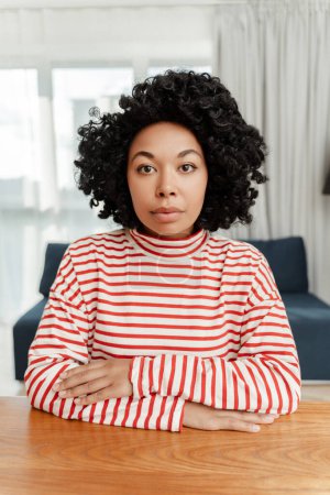 Foto de Retrato de una seria y pensativa mujer afroamericana rizada sentada en el escritorio mirando a la cámara, de cerca. Atractiva mujer auténtica posando para la imagen en la sala de estar - Imagen libre de derechos
