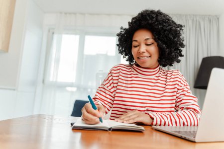 Photo for Portrait of smiling positive female worker sitting at workplace using laptop, making notes in notebook at home. Attractive curly female writer in casual clothes. Project planning, online education - Royalty Free Image