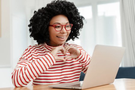 Foto de Sonriendo mujer afroamericana, influencer usando el ordenador portátil, mostrando el gesto del corazón teniendo videollamada - Imagen libre de derechos
