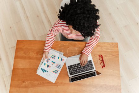 Téléchargez les photos : Portrait d'une femme d'affaires afro-américaine frisée et intelligente assise au bureau, utilisant un ordinateur portable, travaillant avec des documents, des graphiques, une vue d'ensemble. Femme pigiste travaillant en ligne sur le lieu de travail - en image libre de droit