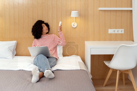 Téléchargez les photos : Portrait d'une belle femme afro-américaine sérieuse bouclée tenant la télécommande, allumant le climatiseur, couchée dans un lit confortable à l'hôtel, utilisant un ordinateur portable, travaillant en ligne - en image libre de droit
