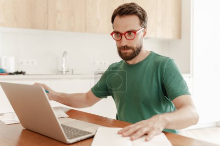 Téléchargez les photos : Portrait d'un étudiant masculin coûteux portant des lunettes rouges et un t-shirt vert décontracté à l'aide d'un ordinateur portable, se préparant à l'examen assis dans la cuisine à la maison. Beau pigiste travaillant avec des documents. Travail à distance - en image libre de droit