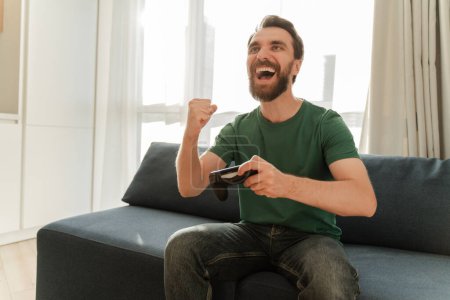 Photo for Attractive man wearing casual clothes sitting on comfortable sofa in apartment playing video games, making winner gesture, copy space. Excited online gamer having fun, leisure time. Technology concept - Royalty Free Image