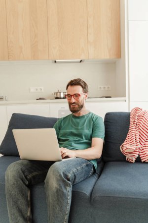Photo for Portrait of positive freelancer working from home, using laptop, sitting on comfortable sofa, online shopping. Attractive young hipster working online in kitchen. ?oncept of technology, remote work - Royalty Free Image