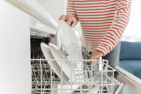 Téléchargez les photos : Femme chargeant la vaisselle dans le lave-vaisselle, plaçant des assiettes, gros plan, foyer de sélection. Nettoyant nettoyage cuisine dans l'appartement. Concept technologique - en image libre de droit