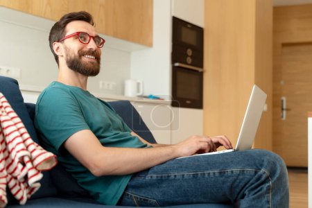 Téléchargez les photos : Homme barbu souriant, programmeur portant des lunettes à l'aide d'un ordinateur portable travaillant en ligne à partir de la maison - en image libre de droit