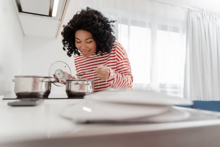 Photo for Portrait of positive African American woman cooking in stylish modern kitchen, holding pan, at home in apartment. Attractive housewife using equipment and dishes. Household concept - Royalty Free Image