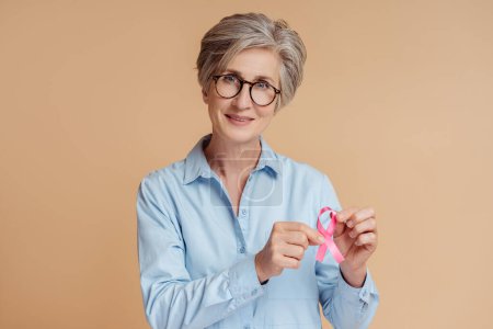 Foto de Mujer mayor confiada sosteniendo cinta rosa aislada sobre fondo beige. Concientización sobre el cáncer de mama - Imagen libre de derechos