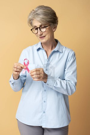 Foto de Mujer mayor de pelo gris sonriente sosteniendo cinta rosa aislada. Mes de sensibilización sobre el cáncer de mama - Imagen libre de derechos