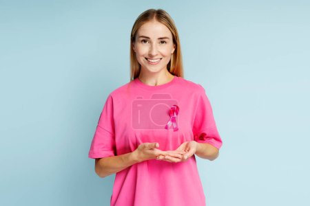 Téléchargez les photos : Femme souriante portant des t-shirts avec un ruban rose cancer du sein isolé sur fond bleu - en image libre de droit