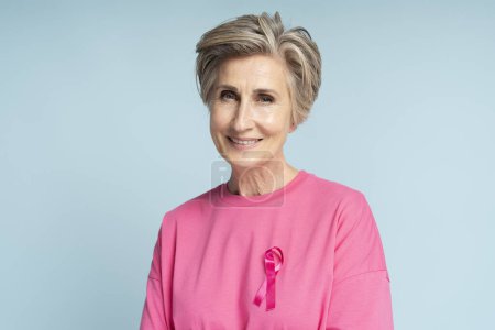 Foto de Mujer mayor sonriente vestida con cinta rosa aislada sobre fondo azul. Concientización sobre el cáncer de mama - Imagen libre de derechos