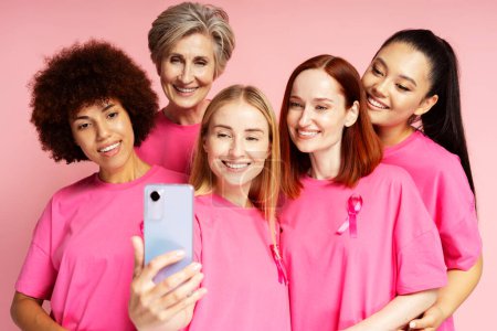 Foto de Mujeres sonrientes con cáncer de mama cinta rosa sosteniendo teléfono móvil aislado sobre fondo rosa - Imagen libre de derechos