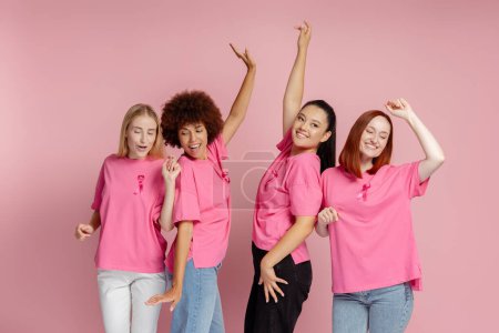 Foto de Mujeres sonrientes y multiétnicas con cinta rosa bailando aisladas sobre fondo rosa. Cáncer de mama - Imagen libre de derechos