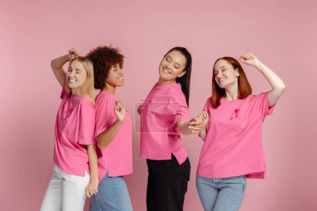 Foto de Grupo de sonrientes mujeres multiétnicas con cinta rosa bailando, celebración aislada sobre fondo rosa - Imagen libre de derechos