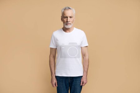 Téléchargez les photos : Homme barbu aux cheveux gris âgé confiant 60-69 ans, retraité adulte âgé vêtu d'un t-shirt blanc et d'un jean bleu décontracté, regardant avec confiance la caméra, isolé sur fond beige - en image libre de droit