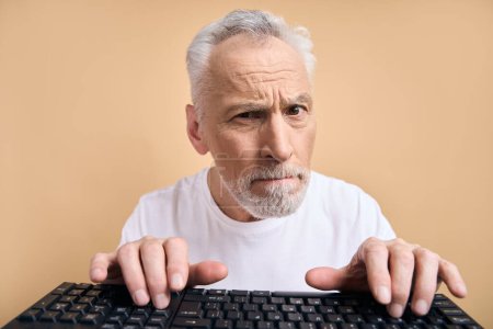 Photo for Excited senior man wearing wearing white t shirt, typing on keyboard shopping online isolated on beige background. Portrait of emotional grey haired programmer working project. Technology concept - Royalty Free Image