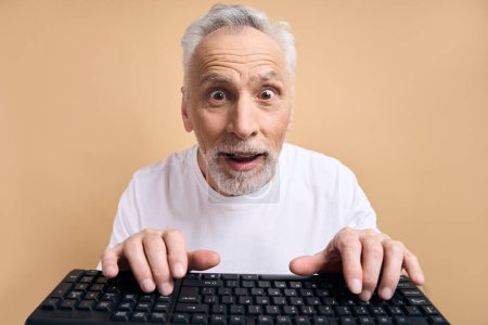 Foto de Emocionado hombre mayor que llevaba puesta camiseta blanca, escribiendo en el teclado de compras en línea aislado en fondo beige. Retrato del proyecto de trabajo del programador de pelo gris emocional. Concepto tecnológico - Imagen libre de derechos