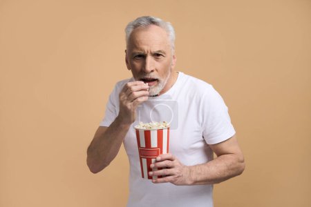 Téléchargez les photos : Incroyable homme âgé barbu aux cheveux gris caucasiens, en t-shirt blanc, regardant attentivement le film et mangeant du pop-corn, exprimant des émotions excitées, isolé sur fond beige. Divertissement des gens Hobby - en image libre de droit