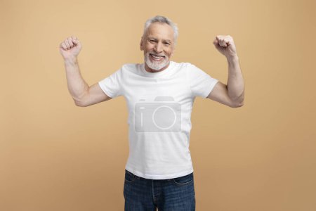 Foto de Hombre mayor sonriente mostrando gesto exitoso aislado sobre fondo beige. Atractivo pensionista posando para la imagen, mostrando los músculos, haciendo ejercicios - Imagen libre de derechos