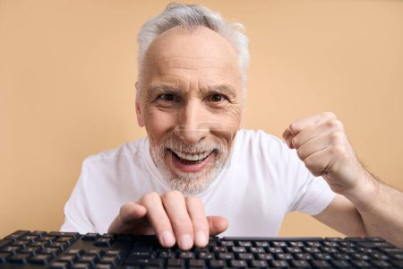 Téléchargez les photos : Homme sénior excité tapant sur le clavier jouant à un jeu vidéo, succès de célébration isolé sur fond beige. Heureux joueur aux cheveux gris émotionnel paris sportifs en regardant la caméra. Concept technologique - en image libre de droit