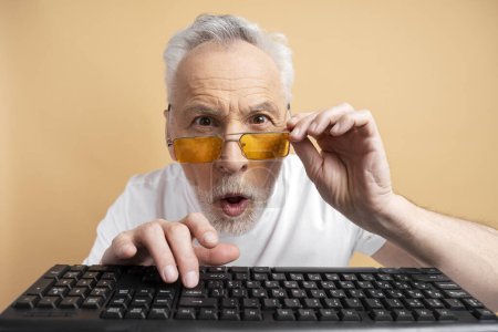 Photo for Surprised mature gray haired man in stylish orange glasses typing on keyboard isolated on beige background. Senior male, business concept - Royalty Free Image