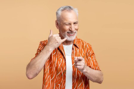 Téléchargez les photos : Poil gris positif senior homme clin d'oeil doigt pointant vers la caméra isolée sur fond beige. Appelez-moi concept - en image libre de droit