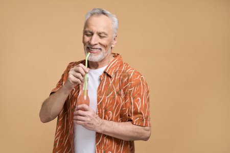 Foto de Caucásico de pelo gris barbudo feliz hombre mayor relajado, con camisa de color naranja brillante, beber una refrescante limonada de una paja, sonriendo, disfrutando de las vacaciones de verano, aislado en el fondo de color beige - Imagen libre de derechos