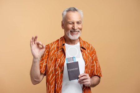 Foto de Retrato de un atractivo hombre de 60 años con una camisa elegante que muestra un gesto okey con pasaporte y una tarjeta de embarque aislada sobre un fondo beige. Concepto de viaje de verano - Imagen libre de derechos