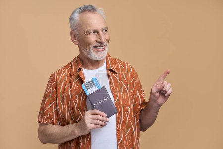 Photo for Happy positive gray haired bearded senior man pointing finger at copy space for ads on beige background, holding a boarding pass, enjoying the upcoming flight. Retired male traveler travelling abroad - Royalty Free Image