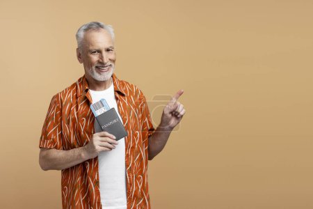Téléchargez les photos : Portrait d'un séduisant homme âgé tenant un passeport et une carte d'embarquement, regardant la caméra, pointant du doigt sur l'espace de copie, mur vide. Concept de voyage - en image libre de droit