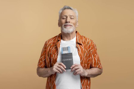 Téléchargez les photos : Portrait d'un homme séduisant de 60 ans les yeux fermés tenant passeport et carte d'embarquement isolés sur fond beige - en image libre de droit
