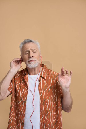 Téléchargez les photos : Homme barbu blanc chevelu gris avec des vêtements décontractés élégants, écoutant de la musique apaisante sur les écouteurs, relaxant avec les yeux fermés sur fond beige isolé. Les gens. Divertissement. Mode de vie - en image libre de droit