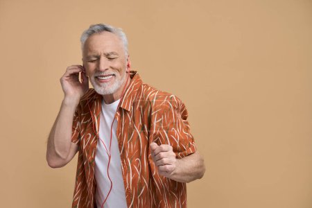 Téléchargez les photos : Élégant moderne caucasien heureux retraité homme âgé, cheveux gris actif touriste masculin voyageur, un joyeux vacancier souriant, profiter des vacances d'été, écouter de la musique sur fond beige isolé - en image libre de droit