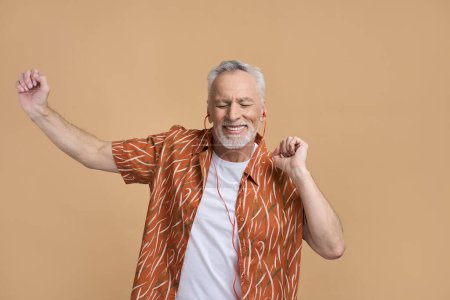 Téléchargez les photos : Blanc élégant homme âgé, heureux retraité fabricant de vacances, portant des écouteurs orange et chemise décontractée, souriant largement, passer à la musique, écouter une bande son cool, sur fond beige isolé - en image libre de droit