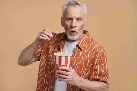 Téléchargez les photos : Portrait d'homme âgé frustré étonné caucasien, retraité mangeant du pop-corn et regardant un film ou une vidéo effrayant, exprimant des émotions effrayées, posant avec la bouche ouverte sur fond beige - en image libre de droit