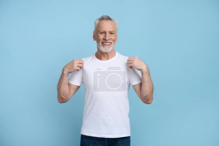 Photo for Smiling Caucasian gray haired bearded senior man showing a white t-shirt he's wearing with mockup, copy space for your ads , smiling looking at camera, isolated over blue background. - Royalty Free Image