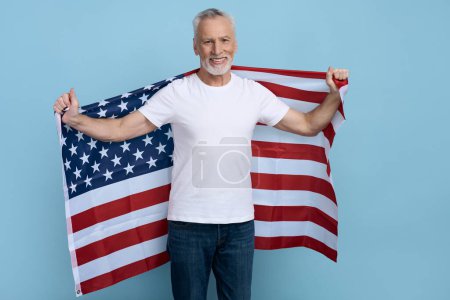 Téléchargez les photos : Beau homme aux cheveux gris et souriant tenant le drapeau américain isolé sur fond bleu. Portrait de joyeux, 60 ans de célébration patriote américain Jour de l'indépendance en regardant la caméra. 4 juillet - en image libre de droit