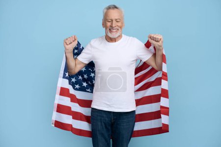 Foto de Hombre mayor feliz en camiseta blanca y vaqueros azules, sonriendo una sonrisa dentada mirando a la cámara, llevando bandera americana, aislado sobre fondo azul. 4 de julio. Concepto del Día del Patriotismo y la Independencia - Imagen libre de derechos