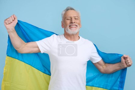 Photo for Happy senior man in white t-shirt, smiling a toothy smile looking at camera, carrying Ukrainian blue and yellow flag, isolated on color background. August 24 . Patriotism and Independence Day concept - Royalty Free Image