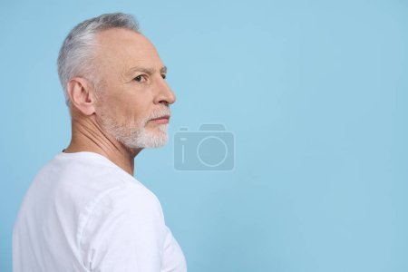 Téléchargez les photos : Portrait latéral d'un homme âgé barbu aux cheveux gris caucasiens, retraité portant un t-shirt blanc décontracté, regardant attentivement de côté, isolé sur fond bleu. Copier l'espace publicitaire. Les gens - en image libre de droit