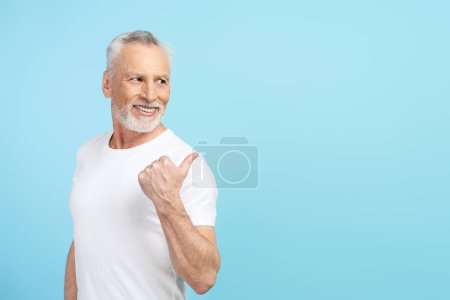 Foto de Hombre sénior positivo con camiseta blanca apuntando con el dedo al espacio vacío, espacio de copia. Atractivo erderly macho mirando hacia otro lado - Imagen libre de derechos
