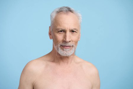 Photo for Confident portrait of a Caucasian gray haired bearded senior handsome man, smiling looking at camera, posing with naked torso on blue background. Healthy lifestyle. Male beauty. Skin and body care - Royalty Free Image