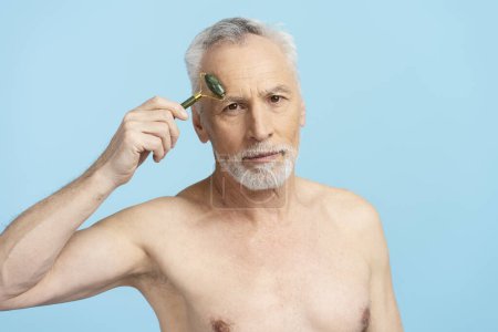 Téléchargez les photos : Portrait d'un homme aux cheveux gris retraité sans chemise faisant un massage facial avec un rouleau facial regardant dans un miroir isolé sur fond bleu. beau senior mâle faire des procédures cosmétiques - en image libre de droit