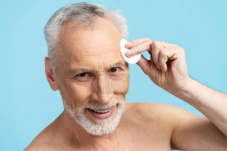 Photo for Closeup of senior man removing makeup with cotton sponge isolated on blue background. Concept of morning routine, skin care - Royalty Free Image