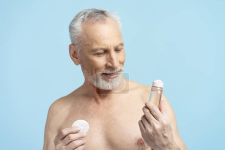 Foto de Hombre de 60 años de edad que aplica agua micelar en el cuidado de la piel sin camisa aislado sobre fondo azul. Concepto de procedimientos de belleza - Imagen libre de derechos