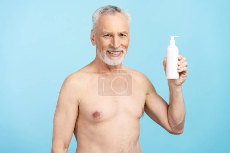 Foto de Retrato de un anciano sonriente con el torso desnudo sosteniendo crema aislada sobre fondo azul. Concepto de cuidado de la piel y del cuerpo - Imagen libre de derechos